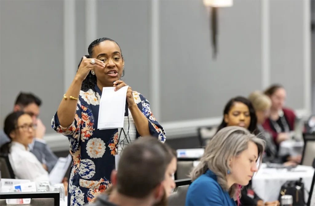 A person is standing an speaking to an audience who are sitting at tables